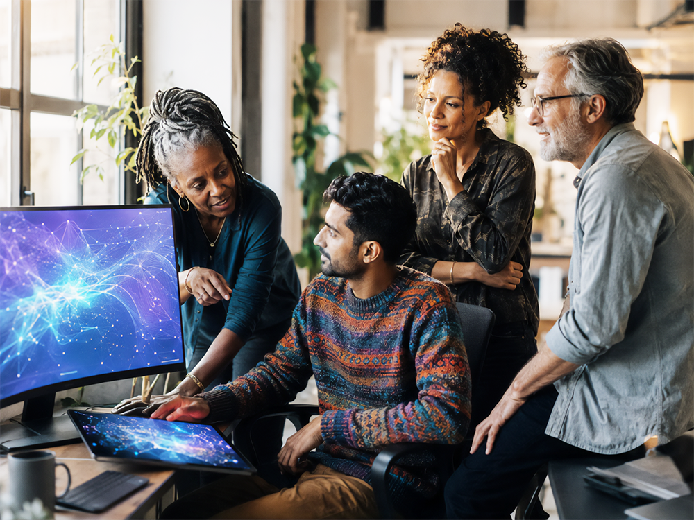 A diverse team of four office colleagues spanning different generations and ethnicities collaborating casually around a curved monitor showing abstract blue and purple neural network patterns inside a sunlit, modern London office workspace.
